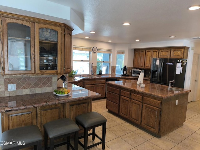 kitchen featuring sink, tasteful backsplash, black fridge with ice dispenser, a center island with sink, and light tile patterned floors