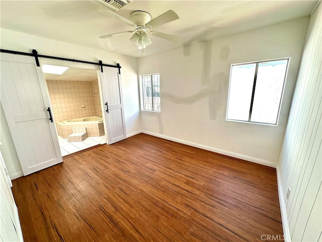 unfurnished bedroom with ceiling fan, a barn door, multiple windows, hardwood / wood-style floors, and connected bathroom