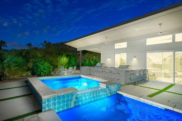 pool at night with an outdoor kitchen, ceiling fan, grilling area, a patio, and an in ground hot tub