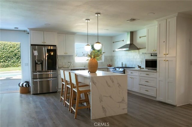 kitchen featuring white cabinetry, wall chimney exhaust hood, stainless steel appliances, decorative light fixtures, and a kitchen island