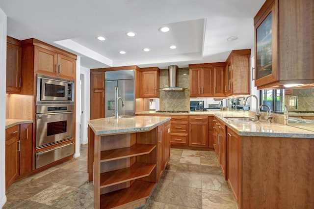 kitchen with decorative backsplash, a kitchen island with sink, wall chimney range hood, light stone counters, and built in appliances