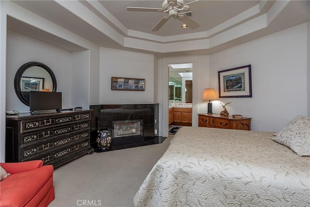 carpeted bedroom with a tray ceiling, ensuite bathroom, a fireplace, and ceiling fan