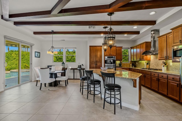 kitchen featuring a notable chandelier, beam ceiling, a kitchen island, wall chimney exhaust hood, and light stone countertops