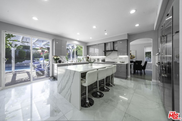 kitchen featuring wall chimney range hood, gray cabinets, light stone countertops, a kitchen island, and a breakfast bar area