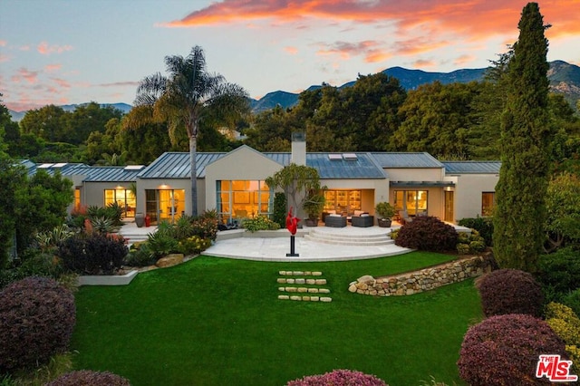 back house at dusk with a mountain view, a lawn, and a patio