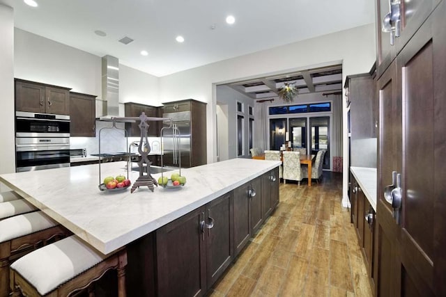 kitchen with a breakfast bar, a kitchen island with sink, wall chimney range hood, appliances with stainless steel finishes, and light hardwood / wood-style floors