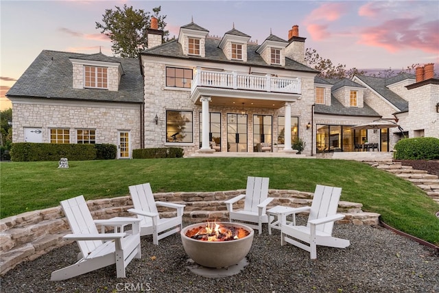 back house at dusk featuring a lawn, a balcony, a patio area, and a fire pit