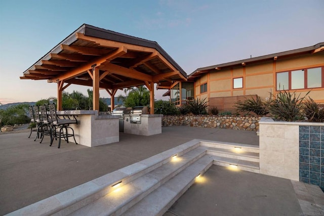 patio terrace at dusk featuring a gazebo, grilling area, exterior bar, and an outdoor kitchen