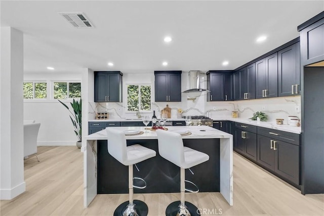 kitchen with backsplash, light hardwood / wood-style flooring, an island with sink, and wall chimney range hood