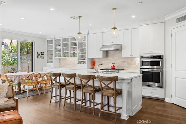 kitchen with hanging light fixtures, stainless steel double oven, dark wood-type flooring, a kitchen island with sink, and white cabinets