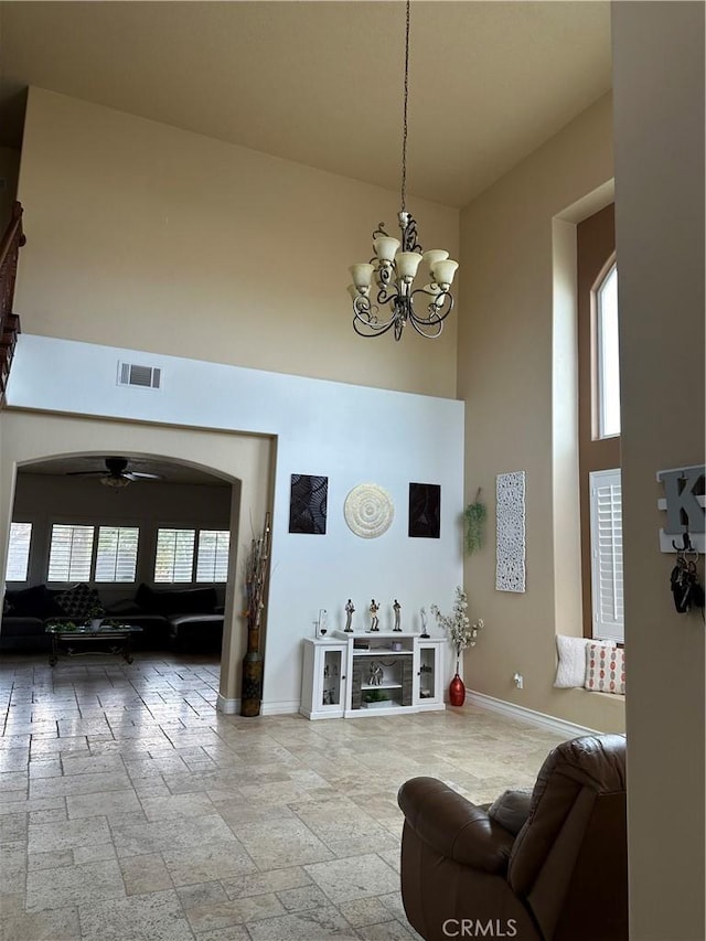 living area with ceiling fan with notable chandelier, a towering ceiling, visible vents, baseboards, and stone tile flooring