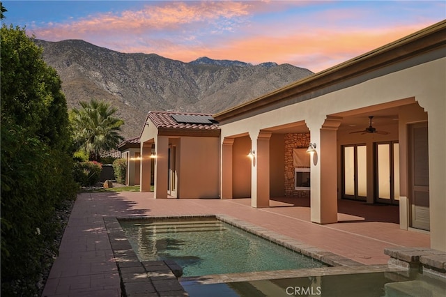 pool at dusk featuring a mountain view, ceiling fan, a patio, and exterior fireplace