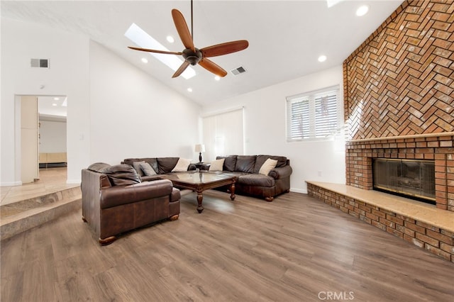 living room with high vaulted ceiling, a brick fireplace, wood-type flooring, ceiling fan, and a skylight