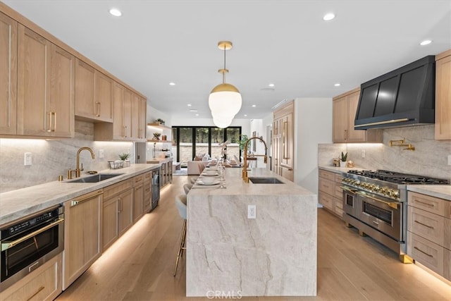 kitchen with wall chimney exhaust hood, decorative light fixtures, stainless steel appliances, sink, and a center island with sink