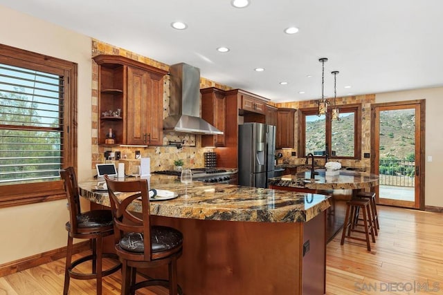 kitchen with stainless steel fridge, decorative light fixtures, wall chimney range hood, dark stone countertops, and light hardwood / wood-style floors