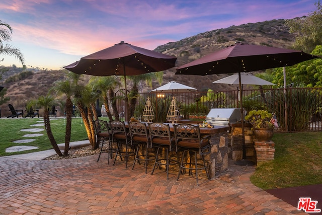 patio terrace at dusk with a mountain view, an outdoor kitchen, area for grilling, and a lawn