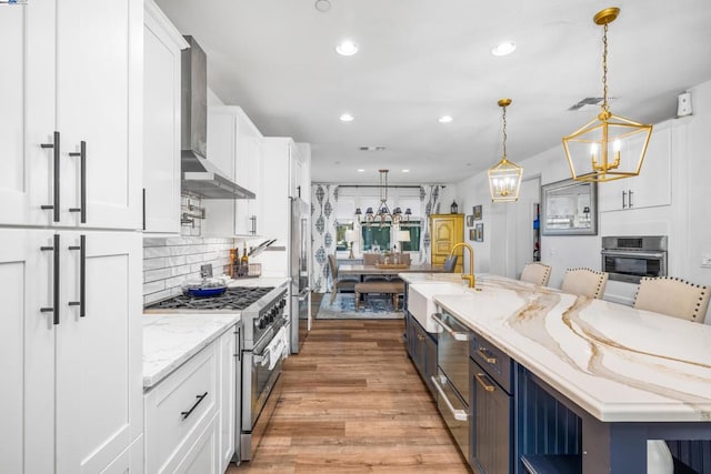 kitchen with premium appliances, wall chimney exhaust hood, hanging light fixtures, and white cabinets