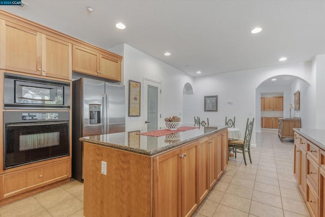 kitchen with dark stone counters, a center island, light tile patterned flooring, black appliances, and light brown cabinetry