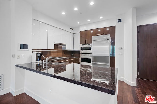 kitchen featuring dark wood-type flooring, kitchen peninsula, dark stone countertops, dark brown cabinets, and appliances with stainless steel finishes