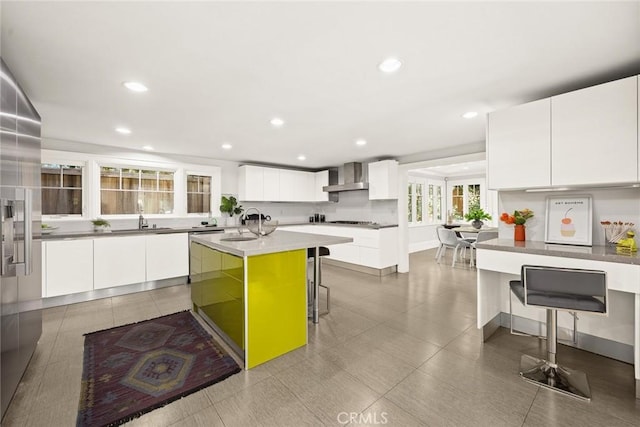 kitchen featuring a kitchen island with sink, sink, wall chimney exhaust hood, white cabinetry, and a breakfast bar area