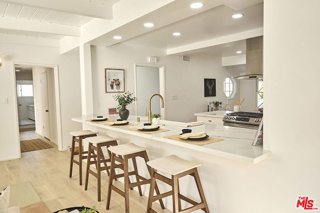 kitchen with a kitchen breakfast bar, wall chimney range hood, stainless steel gas stove, light wood-type flooring, and kitchen peninsula