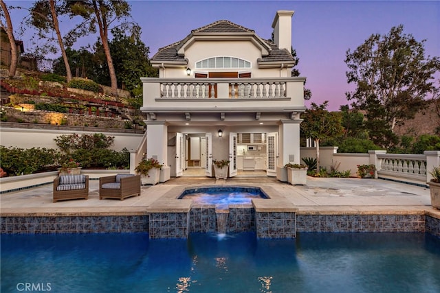 back house at dusk with a swimming pool with hot tub, pool water feature, a balcony, and a patio area