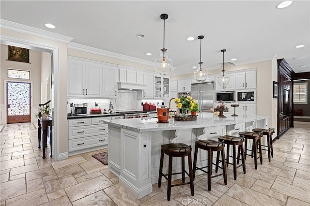 kitchen with hanging light fixtures, built in appliances, a large island with sink, decorative backsplash, and white cabinets