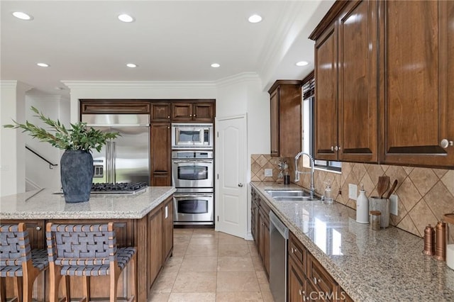 kitchen with sink, light stone counters, built in appliances, crown molding, and a breakfast bar area