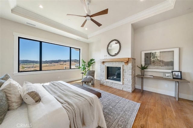 bedroom with light wood-type flooring, a stone fireplace, a tray ceiling, and ceiling fan