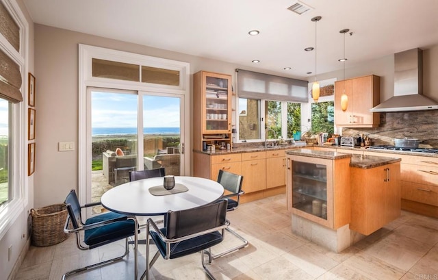 kitchen with light brown cabinetry, wall chimney exhaust hood, decorative light fixtures, and a kitchen island