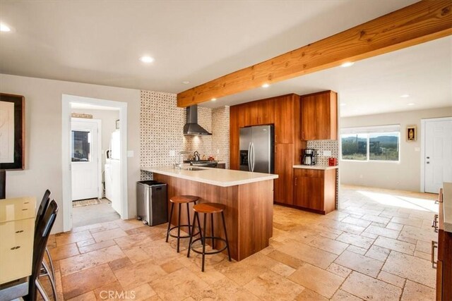 kitchen featuring wall chimney exhaust hood, tasteful backsplash, beamed ceiling, a kitchen bar, and stainless steel fridge with ice dispenser