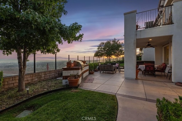 patio terrace at dusk featuring grilling area, a balcony, exterior kitchen, ceiling fan, and an outdoor living space