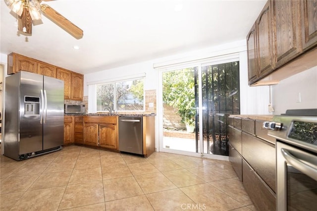 kitchen with ceiling fan, appliances with stainless steel finishes, light stone countertops, light tile patterned flooring, and decorative backsplash