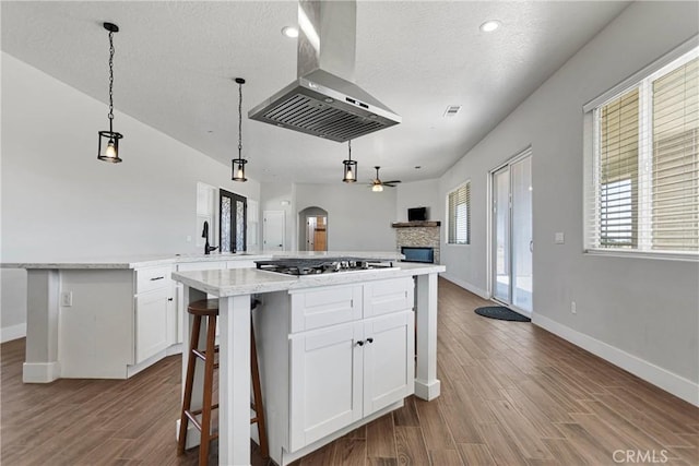 kitchen featuring wall chimney exhaust hood, stainless steel gas cooktop, ceiling fan, a center island, and white cabinetry