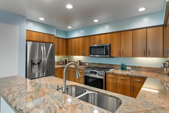 kitchen with light stone countertops, stainless steel appliances, a peninsula, a sink, and brown cabinets