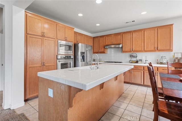 kitchen featuring stainless steel appliances, tile countertops, decorative backsplash, a center island with sink, and light tile patterned floors