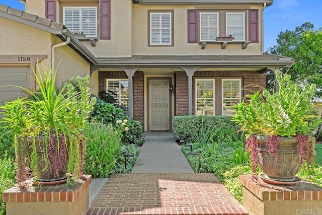 view of exterior entry with a garage, stucco siding, a porch, and brick siding