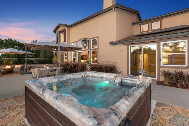 back of property at dusk with a patio, a hot tub, a chimney, and stucco siding