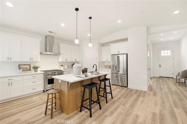 kitchen with high quality appliances, wall chimney range hood, an island with sink, light hardwood / wood-style floors, and white cabinetry