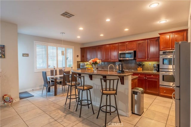 kitchen with stainless steel appliances, a kitchen island with sink, pendant lighting, dark stone countertops, and light tile patterned flooring