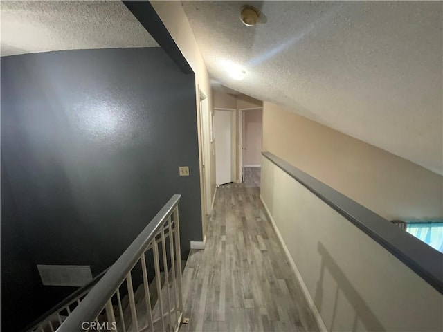 hallway featuring light wood-type flooring, vaulted ceiling, a textured ceiling, and an upstairs landing