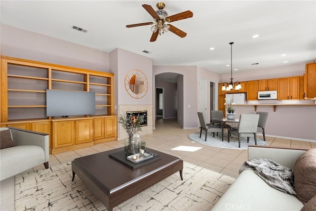 living area with a tiled fireplace, light tile patterned floors, visible vents, arched walkways, and ceiling fan with notable chandelier