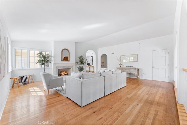 living room featuring arched walkways, a warm lit fireplace, light wood-type flooring, and vaulted ceiling