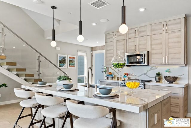 kitchen with appliances with stainless steel finishes, a center island with sink, light brown cabinets, and pendant lighting