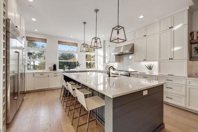 kitchen featuring built in fridge, an island with sink, white cabinetry, light stone countertops, and light wood-type flooring