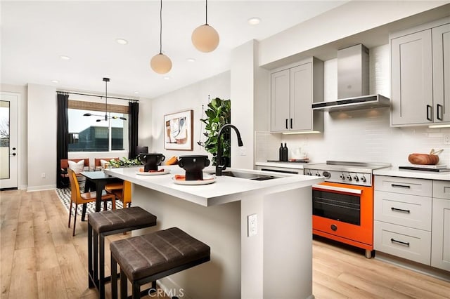 kitchen featuring sink, wall chimney exhaust hood, stainless steel range with electric stovetop, decorative light fixtures, and a center island with sink