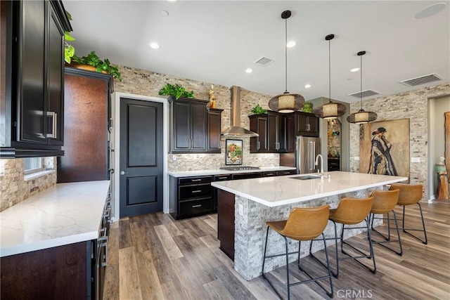 kitchen featuring stainless steel appliances, wall chimney range hood, backsplash, decorative light fixtures, and a kitchen island with sink