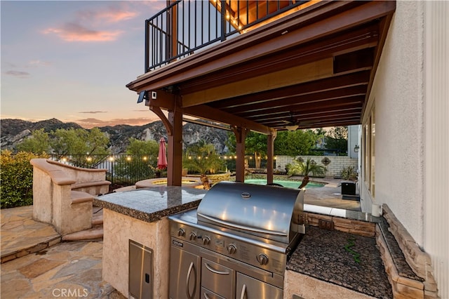 patio terrace at dusk featuring a mountain view, a grill, exterior kitchen, a balcony, and ceiling fan