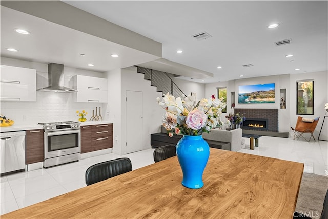 kitchen featuring wall chimney exhaust hood, gas range, white dishwasher, white cabinetry, and a tiled fireplace