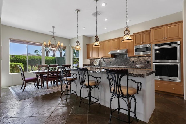 kitchen featuring stainless steel appliances, a notable chandelier, an island with sink, a kitchen bar, and decorative backsplash